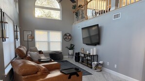 A spacious living room with a leather sofa, a flat-screen TV, a coffee table, and a staircase leading to an upper level. - Coastal Boho Beach Escape - 1 block to paradise (Jacksonville Beach)