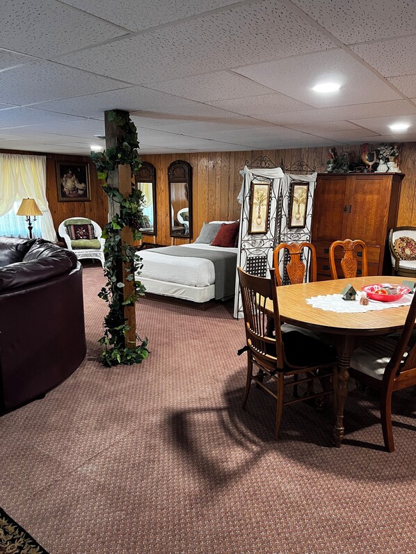 A cozy living room with a wooden floor, a dining table with chairs, a sofa, a bed, and a large mirror.