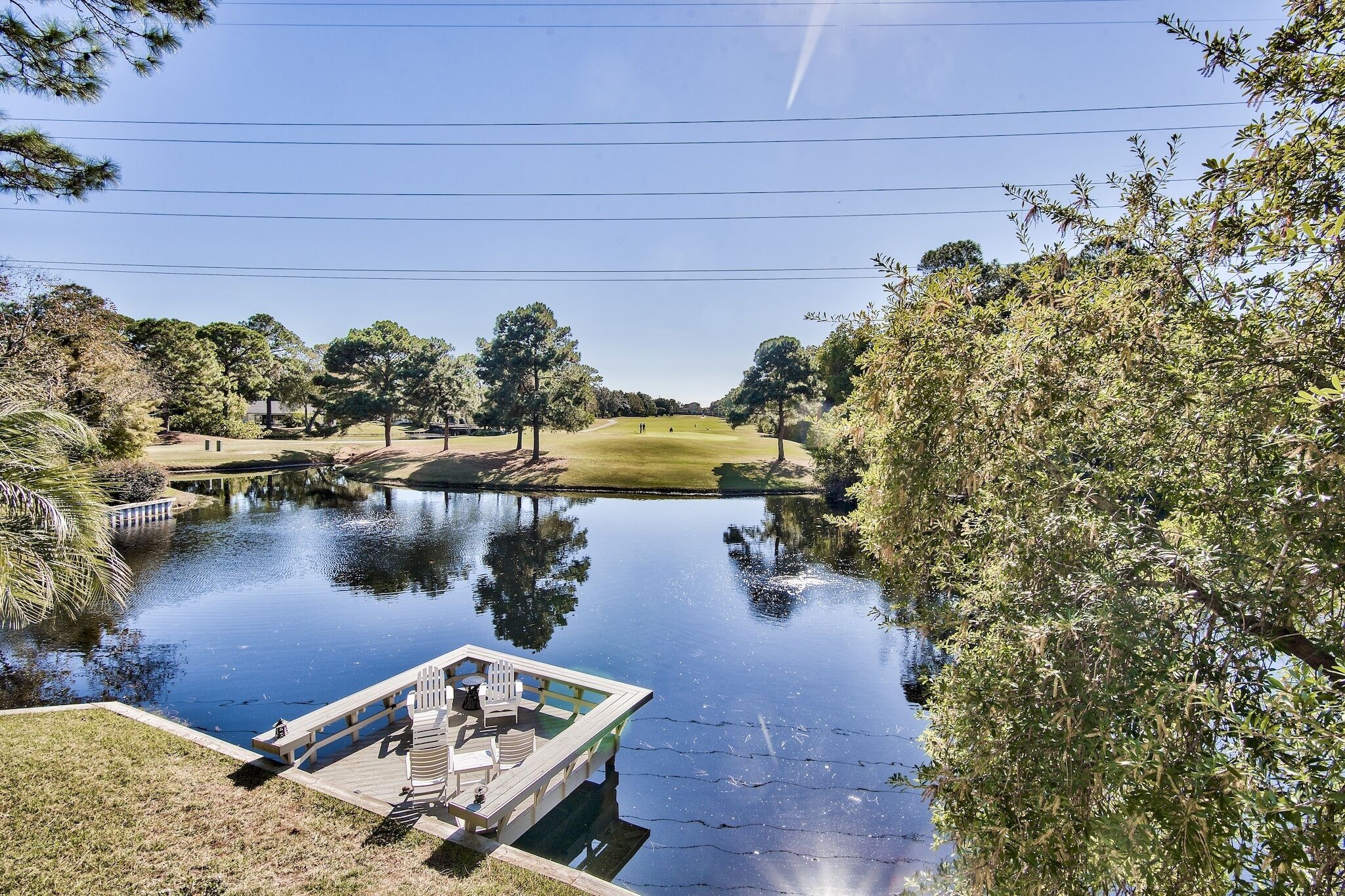 A serene lakeside view with a dock and chairs, surrounded by trees and a golf course in the distance.
