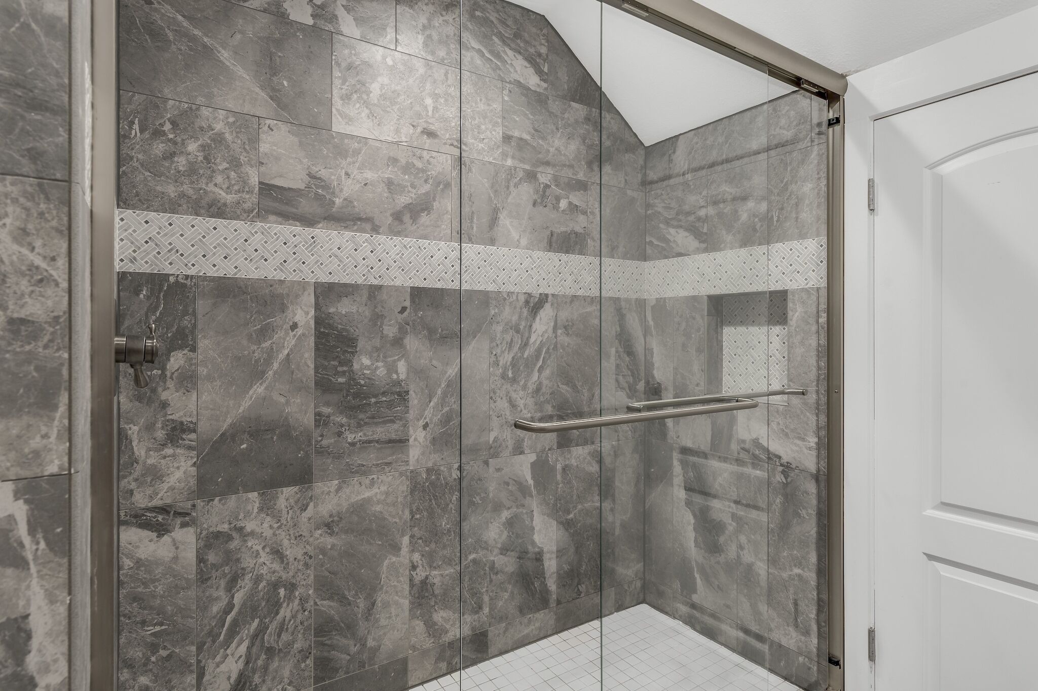 A glass-enclosed shower with a metal handle, grey marble tiles, and a white tiled floor.