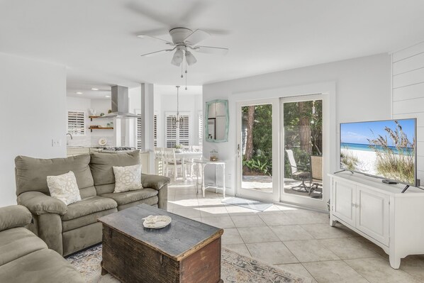A spacious living room with a sofa, coffee table, and a TV on a cabinet. There is a dining area with chairs and a table, and a large window with a view of the outdoors.