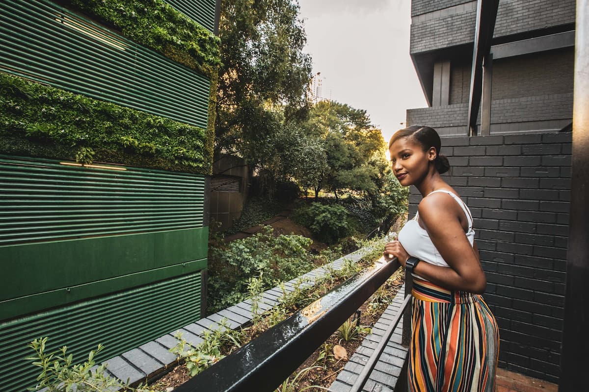 A person standing on a balcony overlooking a garden with a green wall and a brick building in the background.