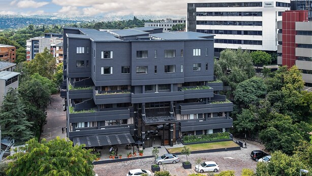 Un immeuble à plusieurs étages doté d’une architecture moderne, avec des balcons et un espace de stationnement où sont garées plusieurs voitures.