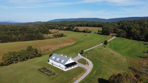 An aerial view of a large, white farmhouse with a red roof, surrounded by expansive green fields and a forested hill in the background. - The Barndominium at Berkeley Ridge - Book your getaway today! (BERKELEY SPRINGS)
