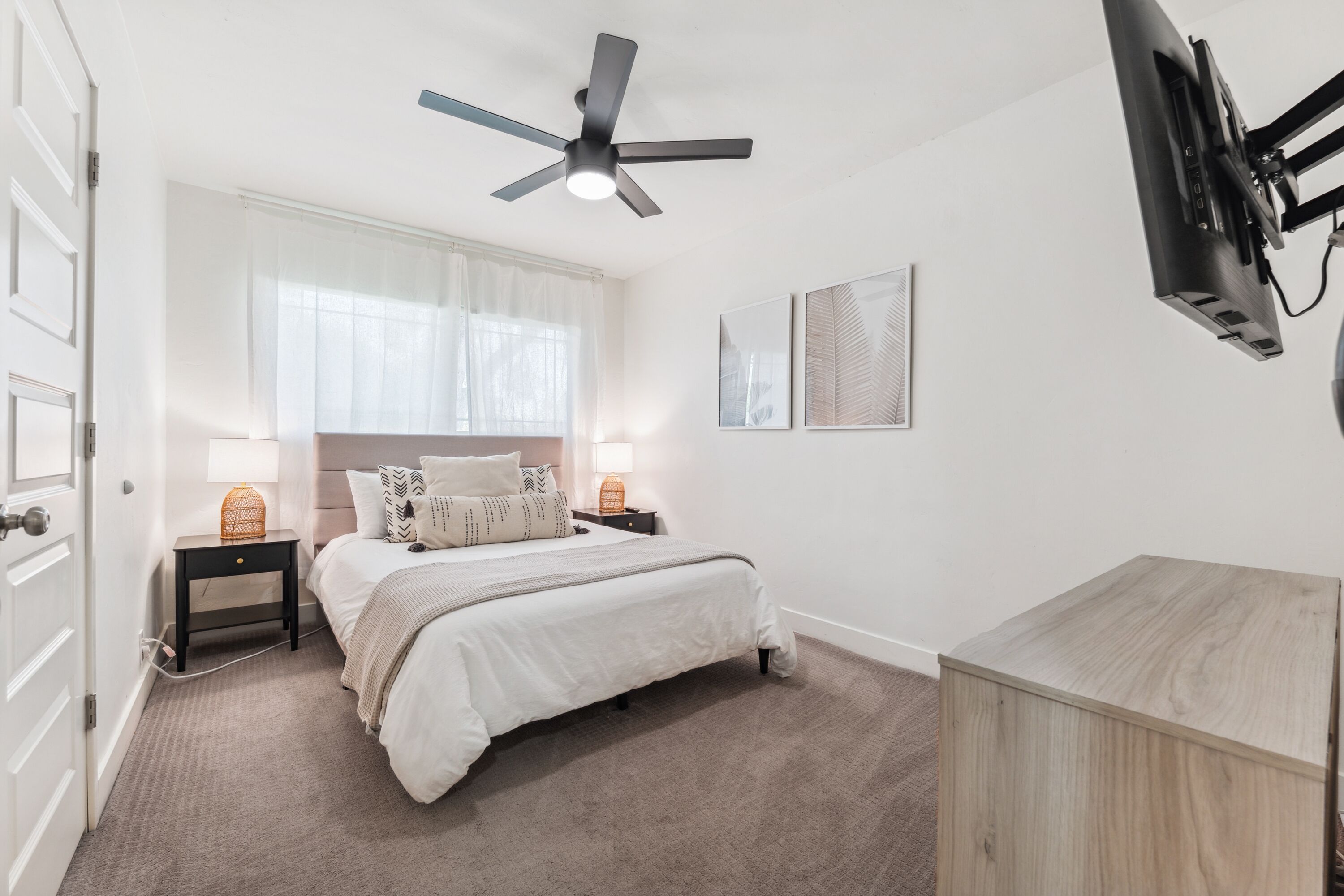 A bedroom with a bed, a ceiling fan, a TV mounted on the wall, and a bedside table with a lamp.