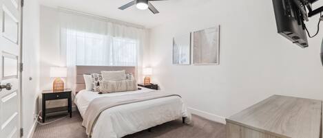 A bedroom with a bed, a ceiling fan, a TV mounted on the wall, and a bedside table with a lamp.