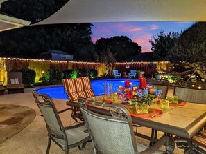 An outdoor patio with a pool, dining table, chairs, and string lights.