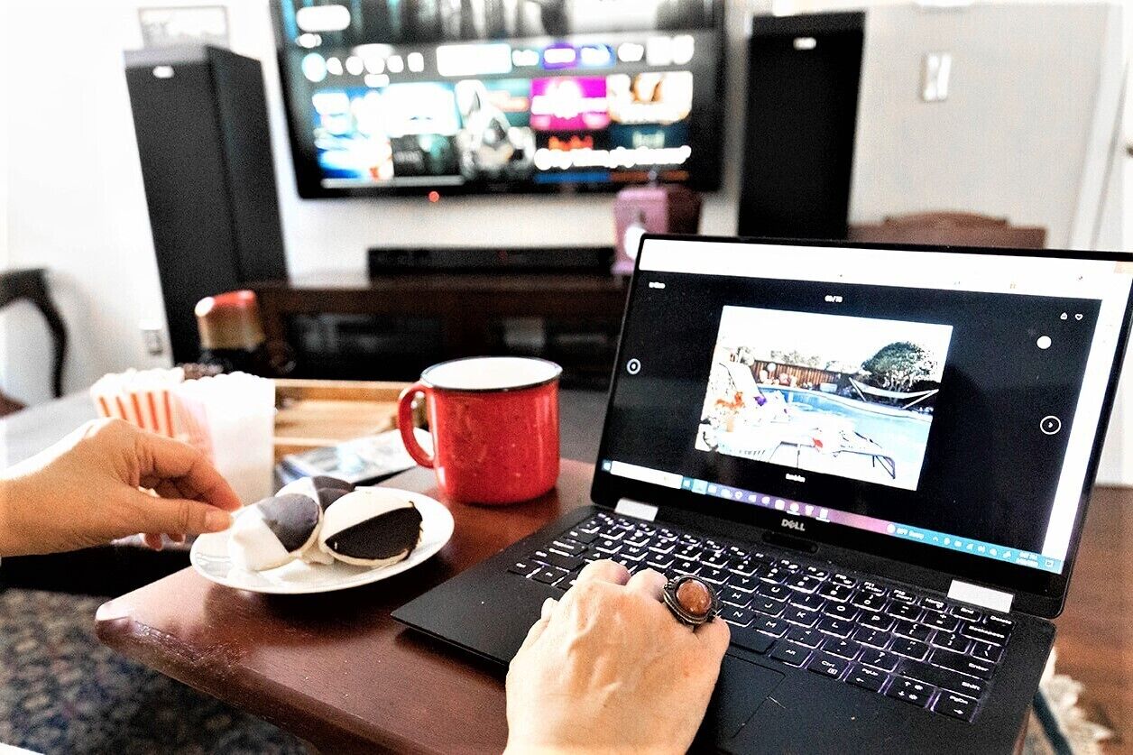 Un ordinateur portable est posé sur une table, une personne l’utilise. On voit également une tasse rouge et une assiette avec de la nourriture.