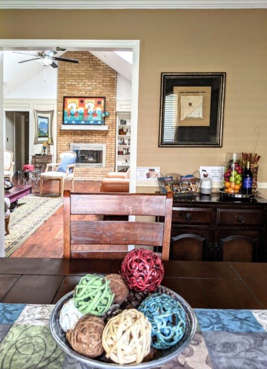 A dining area with a wooden table, a bowl of colorful woven balls, a brick fireplace, and a ceiling fan.