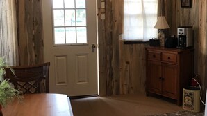A rustic wooden room with a white door, a window with a white curtain, a wooden cabinet, a table, and a lamp. - Cute Farmhouse Cottage  (Oakley)