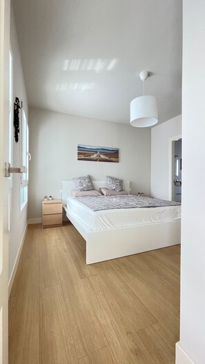 A bedroom with a white bed, a bedside table with a lamp, a painting on the wall, and a wooden floor.