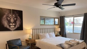 Une chambre d’hôtel avec un lit, une chaise, un ventilateur de plafond, une fenêtre avec des rideaux et un tableau représentant un lion.