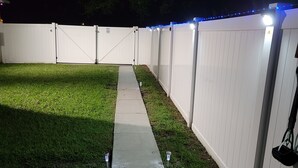 A backyard with a concrete pathway, white fence, and blue string lights.