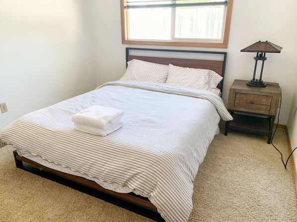 A neatly made bed with a striped bedspread and pillows, a bedside table with a lamp, and a window with blinds. - Peaceful Ranch-style Single level 3B Retreat (Hillsboro)