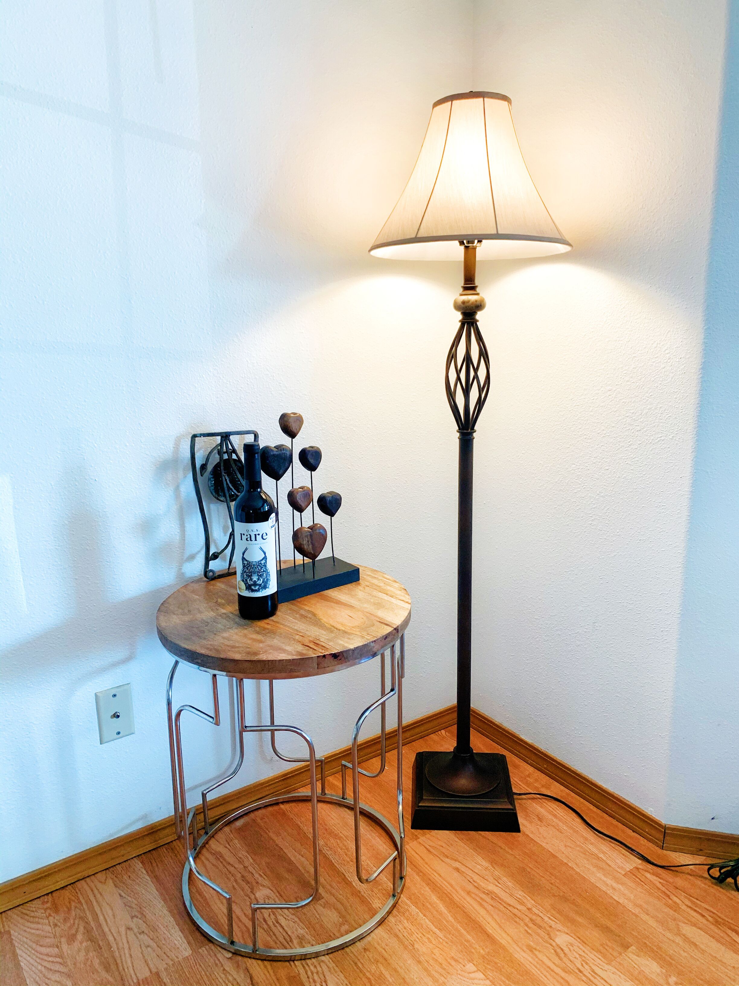 Un rincón con una mesa redonda de madera, una lámpara de pie y un estante en la pared con adornos y una botella.
