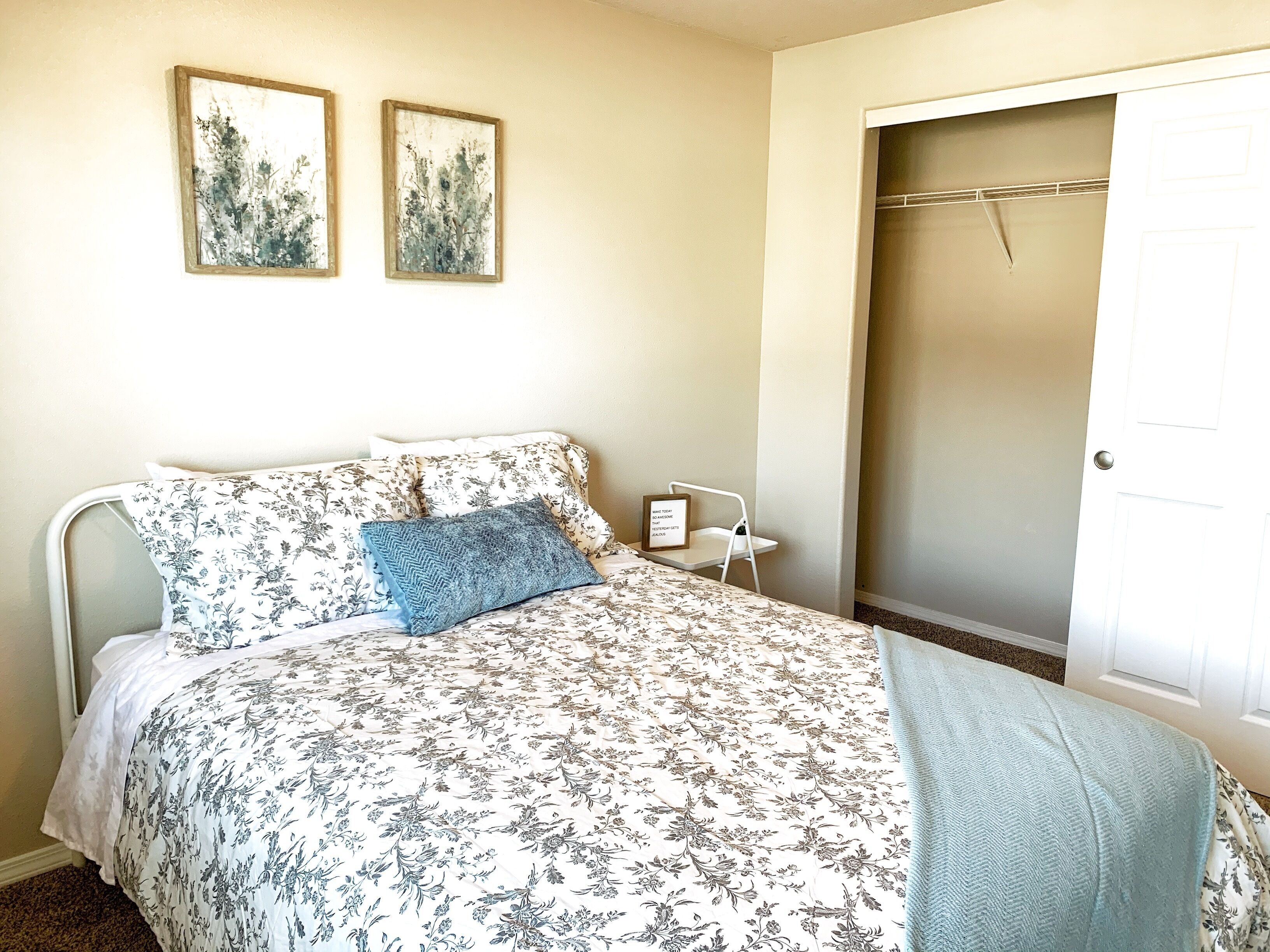 A bedroom with a bed, two wall-mounted paintings, a bedside table with a lamp, and a door.
