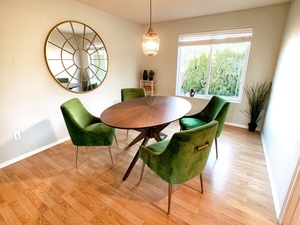 A dining area with a round wooden table surrounded by four green velvet chairs, a large mirror on the wall, and a hanging light fixture.
