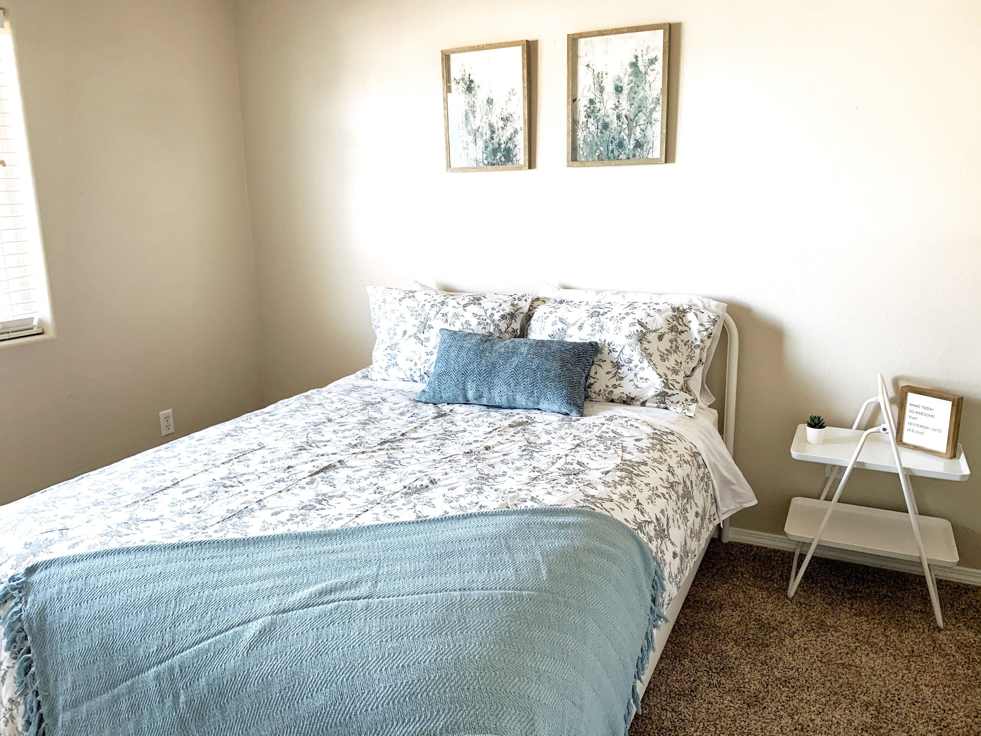 Une chambre à coucher avec un lit, un plaid bleu, une literie à motifs floraux, une petite table de chevet blanche ornée d’une plante, et deux tableaux accrochés au mur.