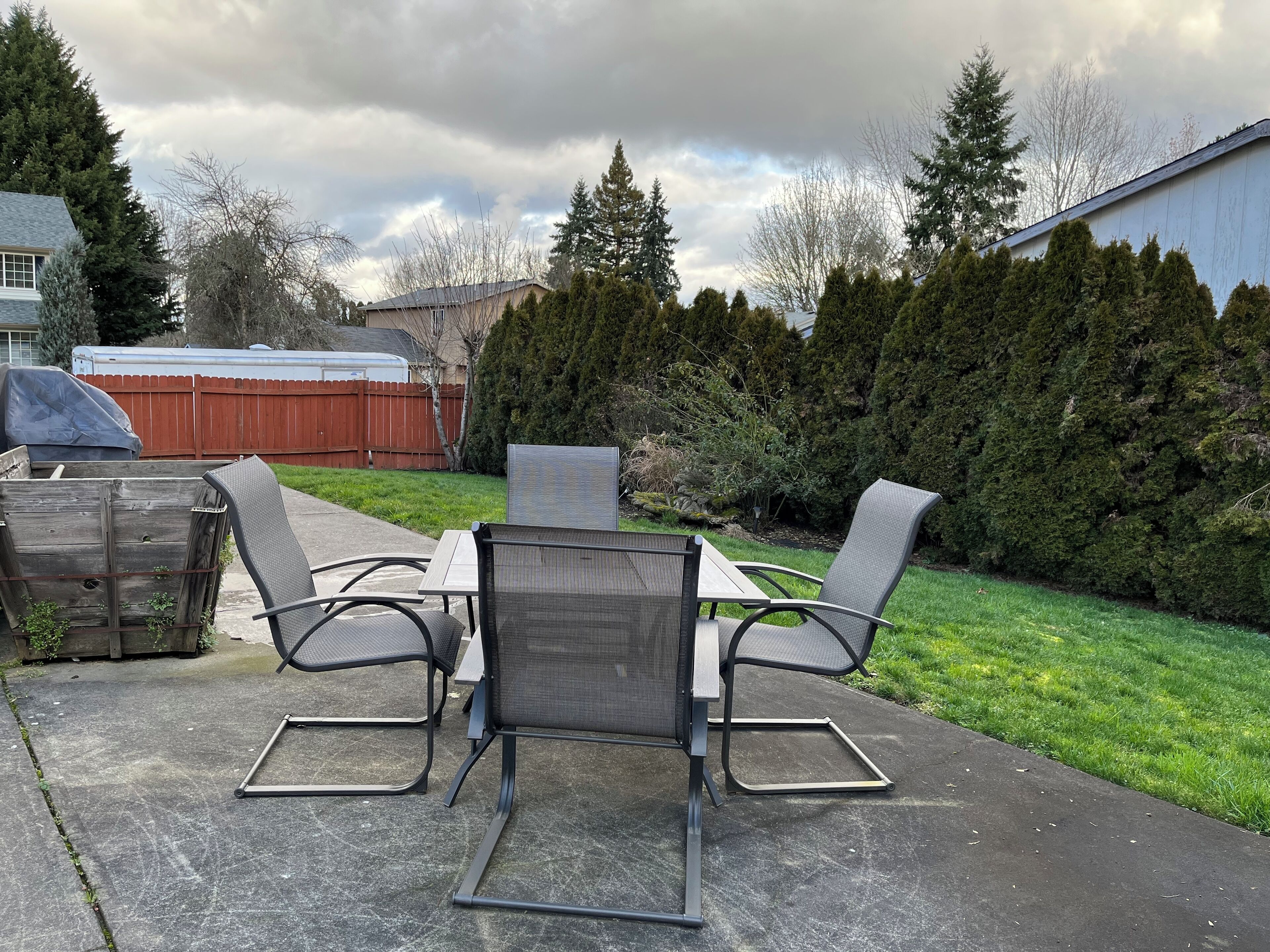 Eine Terrasse mit Tisch und vier Stühlen, ein Holzzaun und eine Reihe immergrüner Bäume.