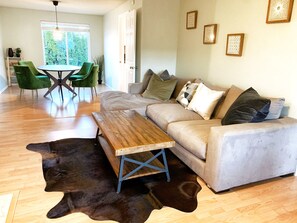 A living room with a sofa, a coffee table, a dining table with chairs, and a large window with a view of greenery.
