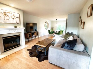 A modern living room with a fireplace, a flat-screen TV, a dining area with green chairs, and a sofa with multiple cushions.