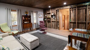 Un salón acogedor con techo de madera, un sillón verde, un reposapiés estampado, una mesa de comedor con manteles individuales a cuadros y una estantería con objetos decorativos.