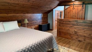 Un dormitorio con techo de madera inclinado, una cama con colcha acolchada, una mesita de noche, una lámpara y una puerta de madera estilo granero.