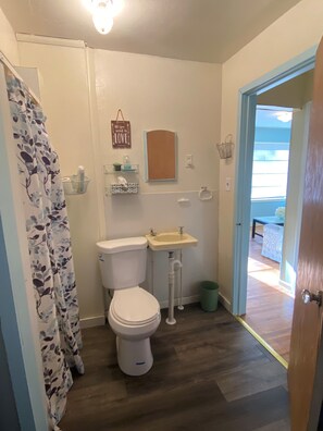 Un baño con inodoro, lavamanos y cortina de ducha. Se ve una ventana con persianas, una puerta y un pequeño estante con artículos de aseo.