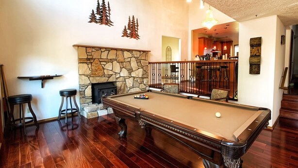 Une table de billard dans une pièce dotée d’une cheminée en pierre, d’un plancher en bois et d’un escalier menant à un niveau supérieur.