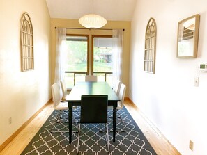 A dining area with a table and chairs, a patterned rug, and a view of greenery outside.