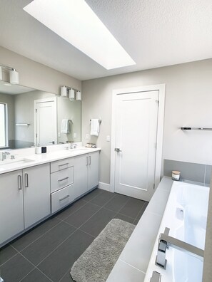 Une salle de bain moderne avec un grand miroir, des meubles blancs, une baignoire et un puits de lumière.