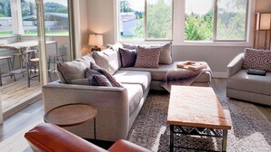 Een moderne woonkamer met een bank, fauteuils, een salontafel en een eethoek met tafel en stoelen. Grote ramen zorgen voor natuurlijk licht en uitzicht op groen.