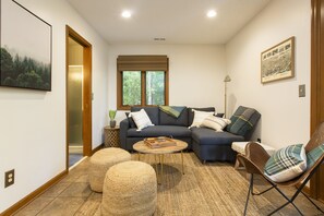 Una sala de estar con un sofá modular, una mesa de centro y una silla. Se ve una ventana con persianas, una lámpara y un cuadro en la pared.