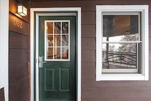 A green door with a window above it, a number '102' on the wall, and a reflection of a fence in the window.