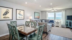 A dining area with a table and chairs, a sofa, a television, and artwork on the walls. - Toes in the Sand 🏖 Drink in Hand!  2 Decks, Views throughout, Great location! (Emerald Isle)