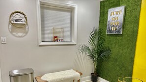 Una habitación con un banco, un basurero, una ventana con persianas, un letrero en la pared, una planta en maceta y una mesita.