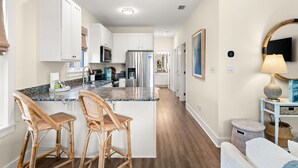 Eine moderne Küche mit weißen Schränken, Edelstahlgeräten und einer Arbeitsplatte aus Granit. Zwei Weidekorbstühle und ein kleiner runder Tisch mit einer Lampe sind ebenfalls vorhanden.