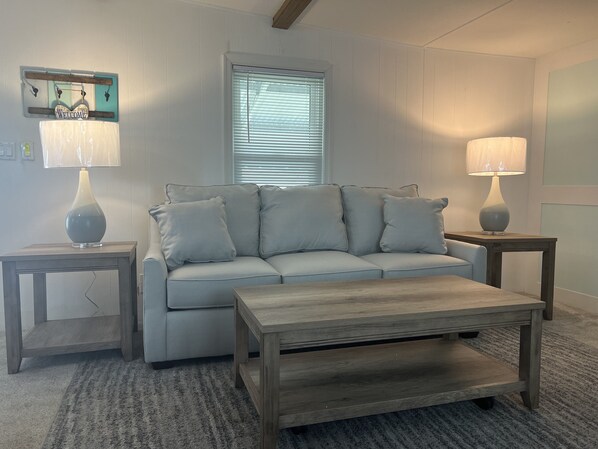 A living room with a light grey sofa, two side tables with lamps, a wooden coffee table, and a window with blinds.
