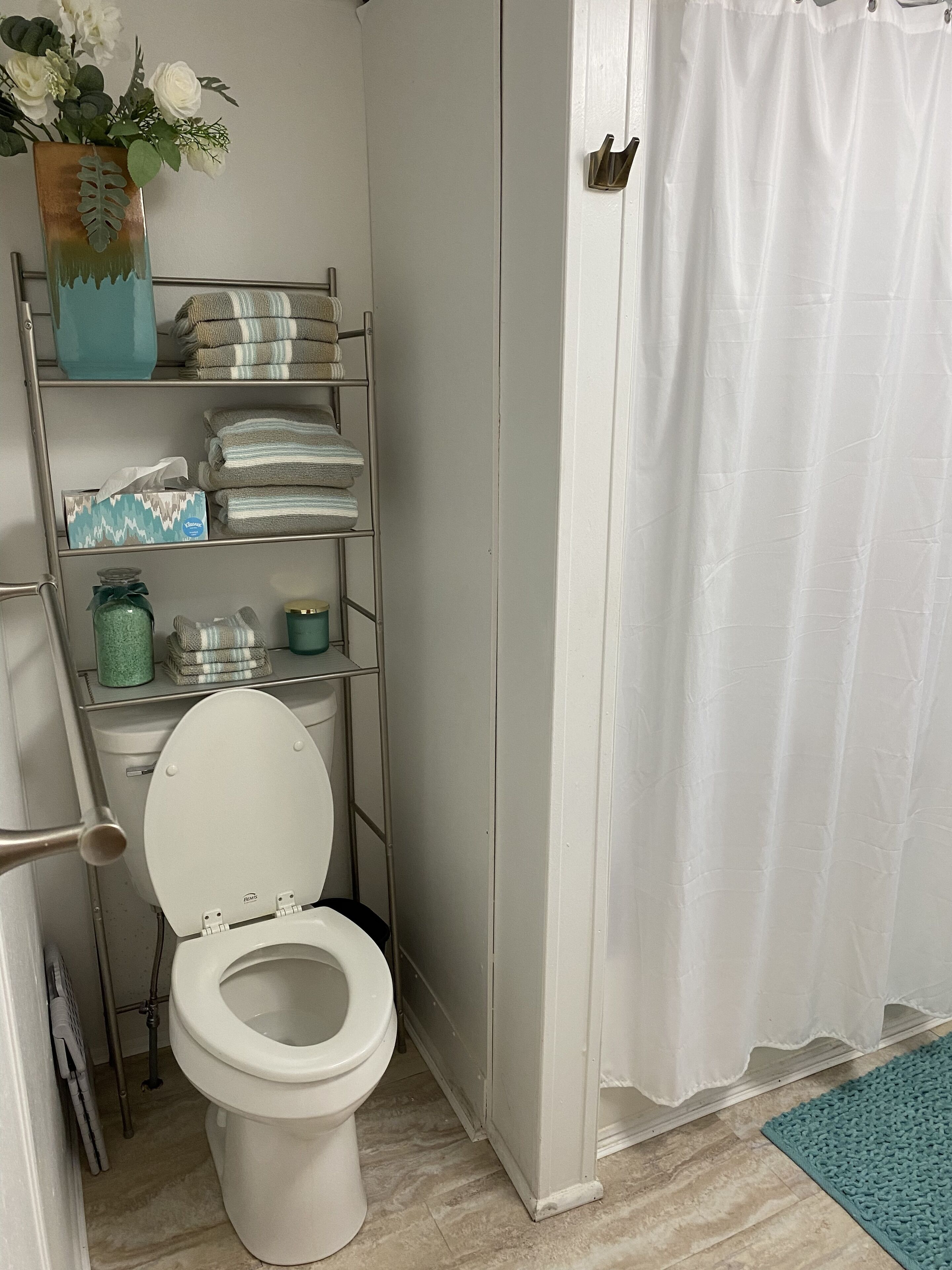 Une salle de bain avec des toilettes blanches, une étagère en métal avec des serviettes et des articles de toilette, un rideau de douche blanc et un tapis bleu sur le sol.
