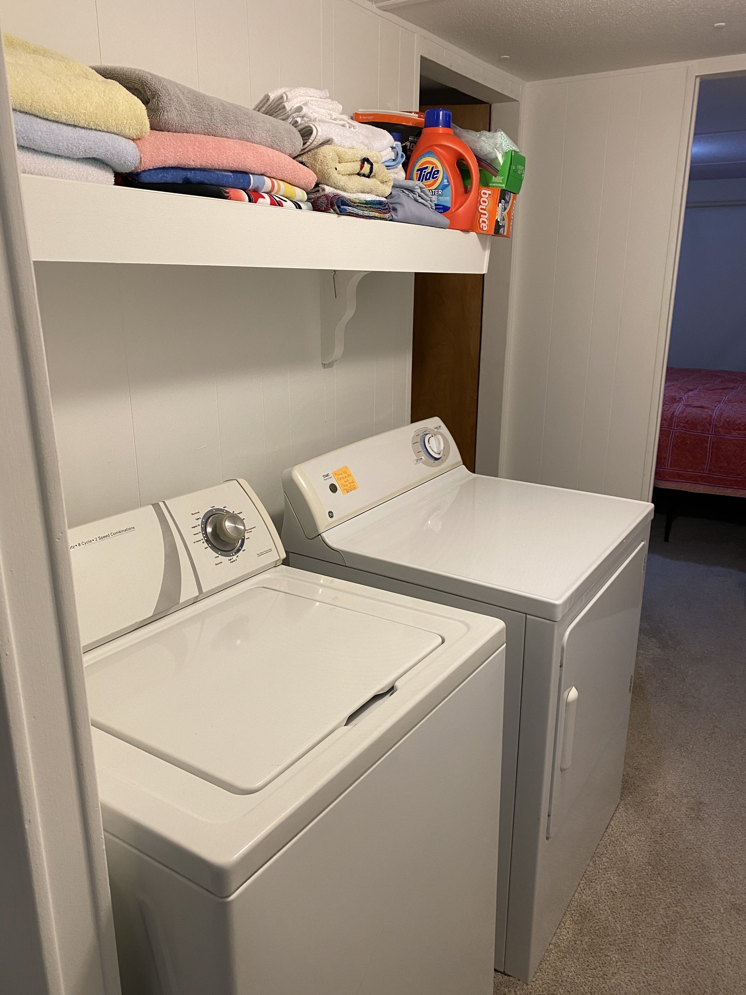 Un espace buanderie comprenant un lave-linge et un sèche-linge à chargement frontal, une étagère avec des vêtements pliés et une bouteille de lessive Tide.