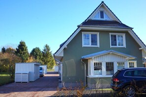 Une maison à deux étages, avec un toit à pignon et un porche à l’avant. On y trouve deux petits abris de jardin et une voiture garée devant.