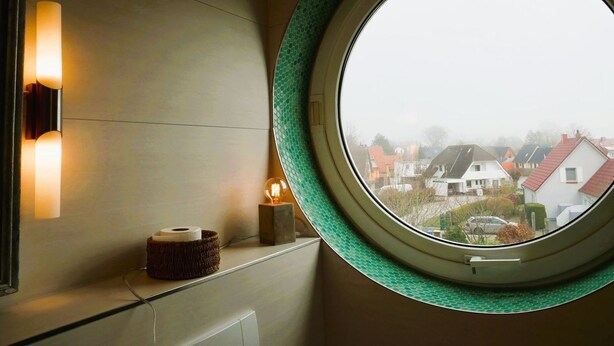 A round window with a green frame, a wall-mounted light fixture, and a small table with a roll of paper and a glass bulb lamp.