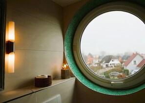 Een rond raam met een groene lijst, een wandlamp en een kleine tafel met een rol papier en een lamp met een glazen bol.
