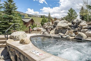 A hot spring with a 33-inch depth warning sign, surrounded by large rocks and pine trees.