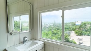 A bathroom with a white vanity, a mirror, and a window offering a view of greenery and a distant building. - Honeymoon Suite (Elbow Cay)