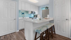 A kitchen with white cabinets, a central island with stools, a microwave, and a tiled backsplash. - Seahorse 512- Seahorse Condos (Gulf Shores)