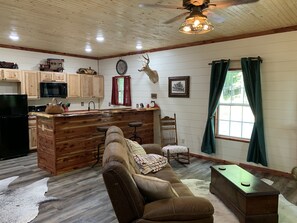 A rustic interior with wooden walls, a kitchen area, a bar, a leather sofa, and a deer head mounted on the wall.