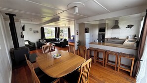 A modern kitchen with a central island, wooden dining table with chairs, and a living area with a sofa and a coffee table. - The Bach on Dorset 🌊 (Opunake)