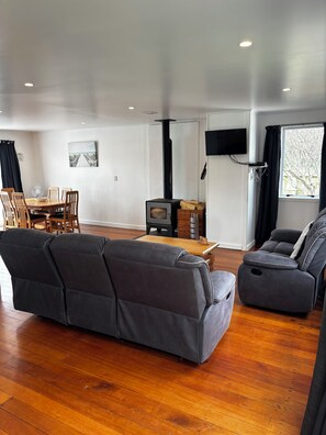 A living room with a sofa, a dining table with chairs, a TV mounted on the wall, and a fireplace. - The Bach on Dorset  🌊 (Opunake)
