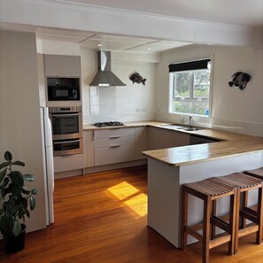 A modern kitchen with a central island, wooden bar stools, a built-in microwave, oven, and a gas stove. - The Bach on Dorset 🌊 (Opunake)